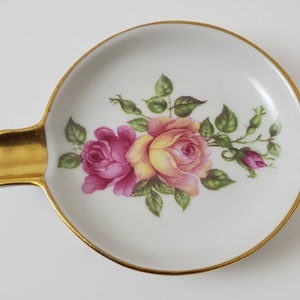 May include: A white ceramic ashtray with a gold rim and a floral design featuring pink and yellow roses.