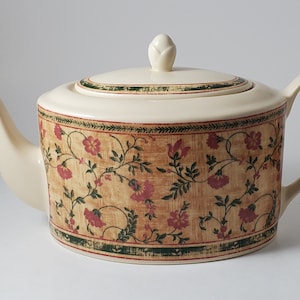 May include: A cream-colored teapot with a floral pattern in shades of red, green, and brown. The teapot has a lid with a knob in the shape of a flower bud.