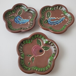 May include: Three small, brown ceramic dishes with scalloped edges. Each dish has a different painted design: a blue grape cluster, a pink flower, and another blue grape cluster.