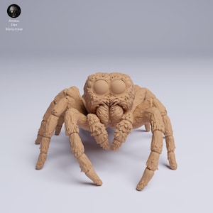 Puede incluir: Miniatura detallada de una ara&ntilde;a saltarina de color beige. El modelo presenta ojos grandes y redondos y un cuerpo y patas texturizados. El logotipo "Animal Den Miniatures" es visible en la esquina superior izquierda.