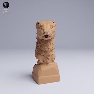 May include: A light brown sculpture of a bear's head and neck, with detailed fur and facial features. The sculpture is mounted on a rectangular base. The logo "Animal des Miniatures" is in the upper left corner.