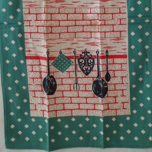 May include: A green tea towel with white diamond pattern border. The center features a red brick wall with black kitchen utensils hanging on hooks.
