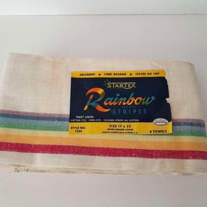 May include: A folded cream-colored towel with rainbow stripes and the text "Rainbow Stripes." The towel is 17 x 33 inches and made of cotton and linen. The label reads "Absorbent, Long Wearing, Leaves No Lint."