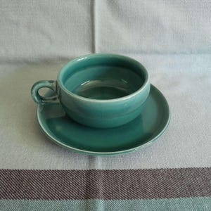May include: A teal ceramic teacup and saucer set. The cup has a rounded shape with a handle, and the saucer is circular. The set is placed on a light-colored fabric with a striped pattern.