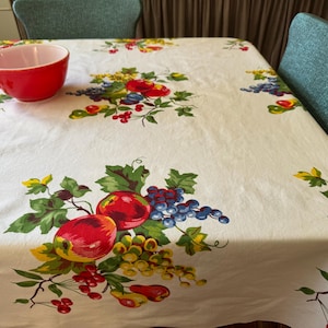 May include: A white tablecloth featuring a repeating pattern of colorful fruit clusters, including red apples, purple grapes, yellow grapes, and green leaves. A solid red bowl sits on the surface of the tablecloth, which is draped over a table.