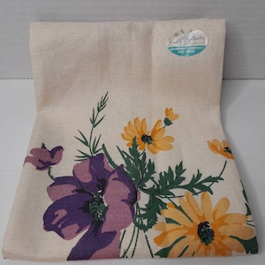 May include: A white tea towel with a floral design featuring purple and yellow flowers. The towel has a label that reads "The Prati of Flanders, Absorbent, Hygienic, Fast Color".