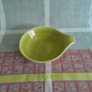 May include: A small, light green ceramic sauce bowl with a handle. The bowl has a smooth, glossy finish and is set on a patterned tablecloth with red, yellow, and white accents. The bowl is empty.