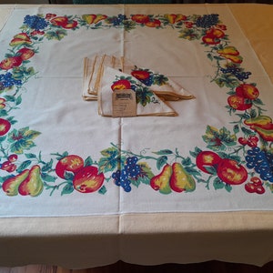May include: A white tablecloth with a yellow border and a fruit pattern. The pattern features pears, apples, grapes, and leaves in shades of red, green, blue, and yellow. A set of napkins is folded on top of the tablecloth.