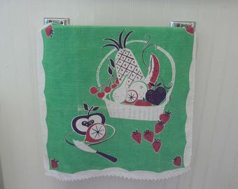 May include: Green kitchen towel with a white scalloped edge, featuring a basket of fruit illustration. The basket includes a pineapple, cherries, and other fruits. A plate with an apple and a knife is also depicted.
