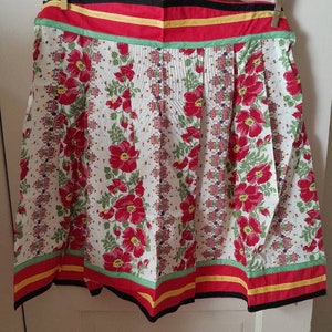 May include: A white apron with a red, yellow, and green striped border. The apron has a floral pattern with red flowers and green leaves on a white background.