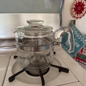 May include: A clear glass stovetop percolator coffee pot featuring a transparent lid, a sturdy glass handle, and a central metal band with the word STAINLESS embossed on the side. The coffee maker sits on a black metal burner grate.