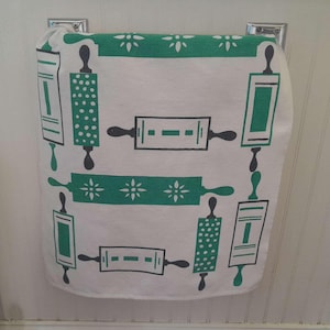 May include: White kitchen towel with a repeating pattern of green rolling pins and floral accents. The towel is hanging on a chrome towel bar. The design is a retro style.