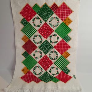May include: White kitchen towel with a patchwork design featuring red, green, and yellow squares with a wreath pattern. The towel has a fringed edge and the text "C.R.C." in the bottom right corner.