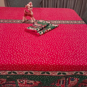 May include: A red tablecloth with white polka dots and a green and white border. The tablecloth has a festive Christmas theme with images of candy canes, holly, and a toy train. A small white ceramic reindeer figurine is sitting on the tablecloth.