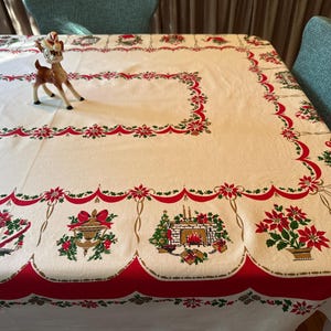 May include: A white rectangular tablecloth featuring a red scalloped border and a festive print of poinsettias, green holly, and Christmas motifs including a fireplace, bells, and an open book with the text NOEL. A small deer figurine stands on top.