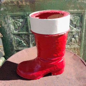 May include: A red boot-shaped planter with a white rim. The planter is made of a glossy material and sits on a rusty metal surface. The background features a green, ornate metal structure.