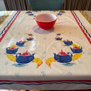 May include: A rectangular tablecloth with a cream-colored base featuring a repeating pattern of blue teapots with red lids on yellow ribbons, bordered by red and blue stripes and yellow diamond accents, with a solid red mixing bowl centered on top.