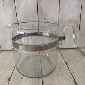 May include: A clear glass coffee carafe featuring a wide, polished silver-tone metal band encircling the upper portion of the body. The vessel includes a sturdy, integrated glass handle and a small pouring spout, showcasing a transparent design.
