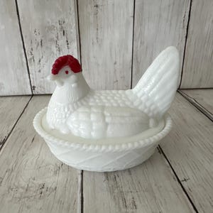 May include: A white milk glass hen on a nest dish featuring a detailed chicken figure with a bright red comb and wattle. The base is designed to resemble a woven basket with a textured rim, while the lid depicts a sitting hen with molded feather details.
