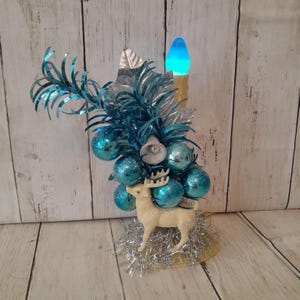 May include: A vintage Christmas decoration featuring a cream-colored reindeer figurine with blue ornaments and tinsel. The decoration includes a blue light-up candle and silver accents, creating a festive holiday display.