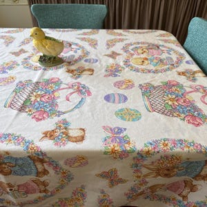 May include: White Easter tablecloth with a repeating pattern of bunnies, baskets of flowers, and Easter eggs. A yellow chick figurine sits on the table. The tablecloth is decorated with pastel colors.