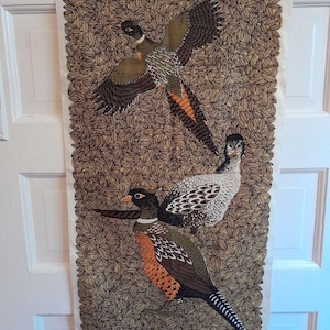 May include: A rectangular fabric wall hanging featuring three stylized pheasant illustrations. The birds are depicted in shades of brown, black, orange, and white against a backdrop of brown leaves. The fabric has a white border.