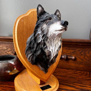 May include: A detailed sculpture of a wolf's head mounted on a wooden plaque. The wolf is depicted with realistic fur in shades of grey and white. The plaque is shield-shaped and attached to a round wooden base. The base has a small black label that reads "WOLF".