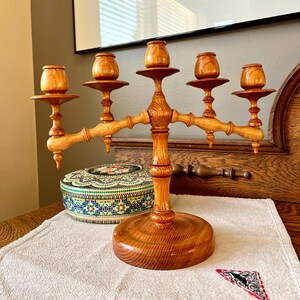 Peut inclure: Un chandelier en bois avec cinq bougeoirs. Le chandelier est en bois de couleur claire et possède une base ronde. Une boîte décorative avec un motif floral et un petit textile triangulaire avec un motif noir et blanc sont également visibles.