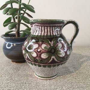 May include: A small, decorative ceramic pitcher with a handle. The pitcher features a green, brown, and white floral design. The pitcher is next to a small blue ceramic pot with a green plant.