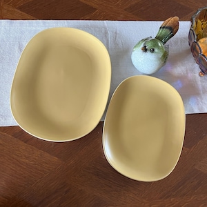 Puede incluir: Dos platos de cerámica de color amarillo mostaza, de forma ovalada. Los platos tienen una superficie lisa y una forma ligeramente irregular. En el fondo se ve una figura decorativa de pájaro con detalles verdes y marrones, junto con un cuenco de cristal decorativo.