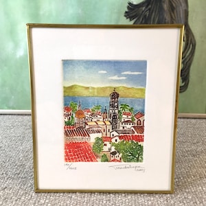 May include: Framed watercolour painting of a coastal town with red rooftops, a church with a bell tower, and a blue body of water in the background. The painting is signed "Madalupe 1995" and numbered "140/1600".