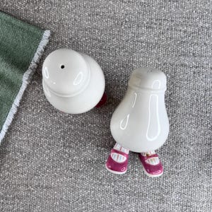May include: A pair of white ceramic salt and pepper shakers shaped like people with pink shoes. The shakers have a glossy finish.