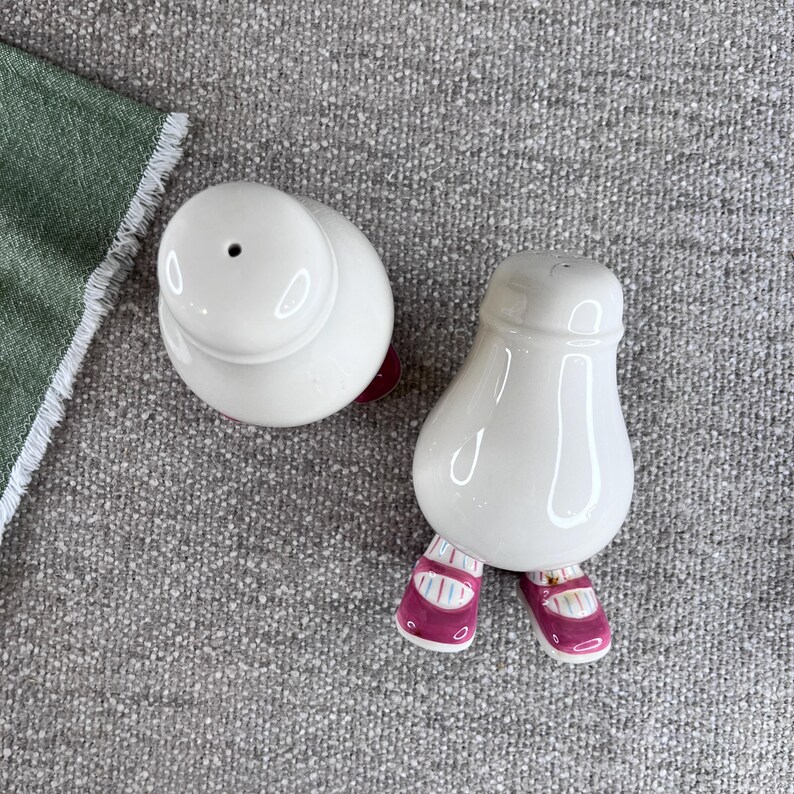 May include: A pair of white ceramic salt and pepper shakers shaped like people with pink shoes. The shakers have a glossy finish.