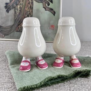 May include: A pair of ceramic salt and pepper shakers shaped like people with pink and white striped shoes. The shakers are white and have a glossy finish.