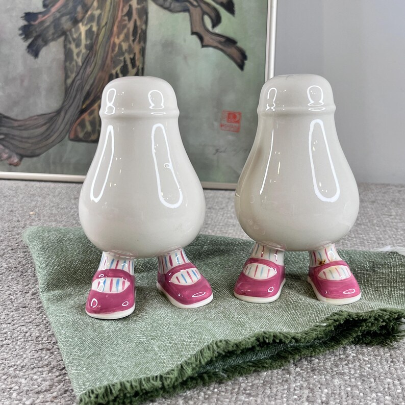 May include: A pair of ceramic salt and pepper shakers shaped like people with pink and white striped shoes. The shakers are white and have a glossy finish.
