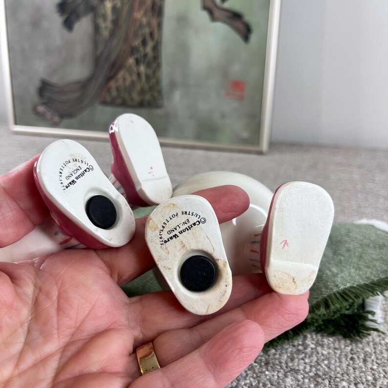 May include: Three white ceramic figurines with pink accents. The figurines are shaped like shoes and have a black magnet on the bottom. The figurines are marked "Carlton Ware, England, Lustre Pottery".