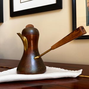 May include: A copper-toned coffee pot with a wooden handle and lid, set on a white cloth. The pot has a brass spout and a slender, angled handle. The design is vintage and elegant.