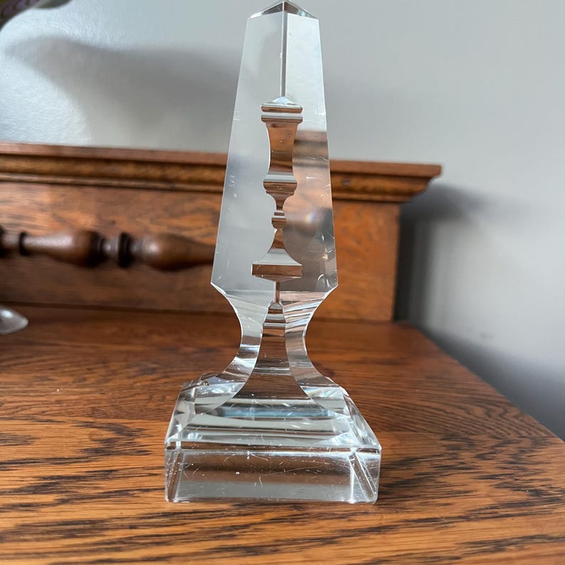 May include: A clear glass obelisk with a decorative design of a stylized vase inside. The obelisk has a square base.