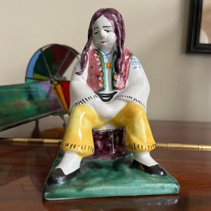 Puede incluir: Una figurita de cerámica de una persona sentada en un taburete. La persona lleva una camisa blanca, pantalones amarillos y un chaleco rojo. La figurita está pintada con detalles en blanco, rojo, amarillo, verde y azul.