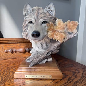May include: A detailed sculpture of a wolf's head, with realistic fur texture and lifelike eyes. The wolf is positioned with a branch and leaves, mounted on a wooden base. The base has a small plaque that reads "SILENT VIGIL".