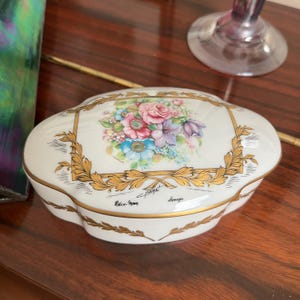 May include: White porcelain trinket box with a gold trim and floral design. The box has a floral design with pink, purple, blue, and yellow flowers. The box is inscribed with "E. Praget Limoges".