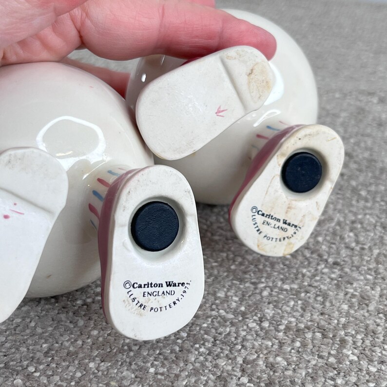 May include: Two white ceramic salt and pepper shakers shaped like people wearing red and white striped shoes. The bottom of each shaker has a black circle and the text "Carlton Ware, England, Lustre Pottery 1975".