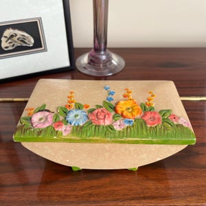 May include: A vintage trinket box with a floral design. The box is made of papier-mâché and features a raised floral design with pink, yellow, orange, blue, and green flowers. The box has a hinged lid and sits on four small feet.