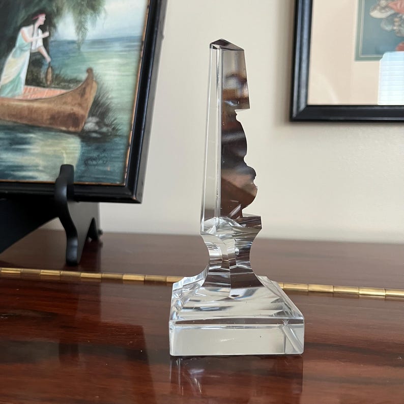 May include: A clear glass bottle stopper with a faceted design. The stopper is shaped like a key and has a square base.