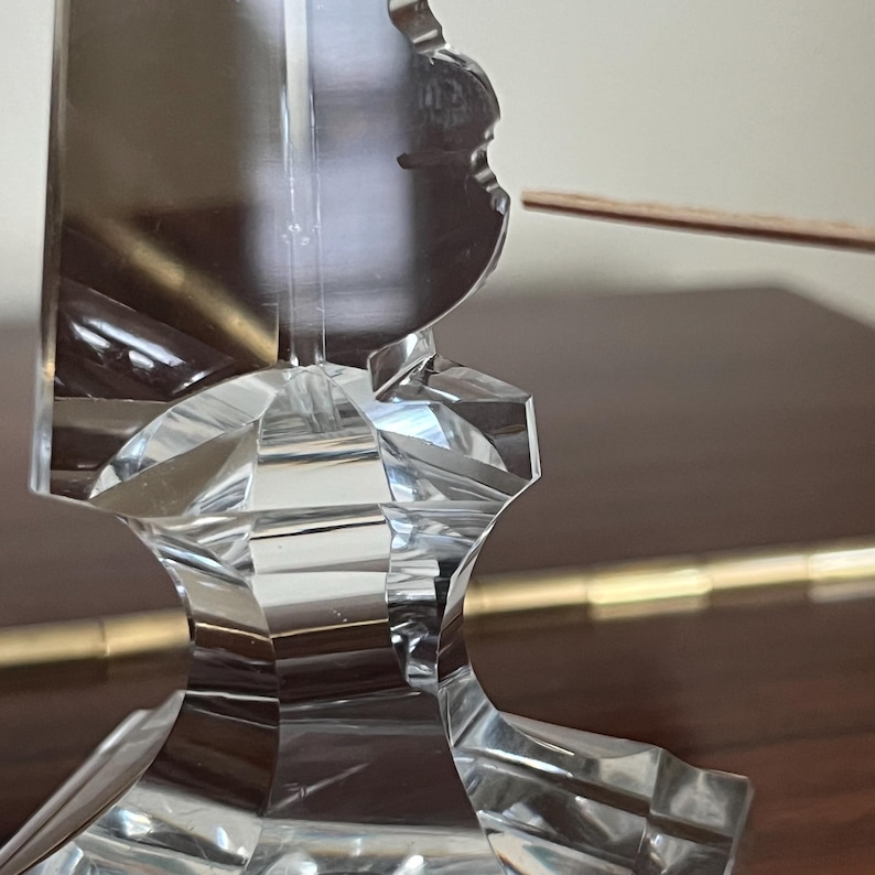 May include: A clear glass crystal candlestick with a geometric design. The base is a square with a raised center and the top is a tall, slender spire.