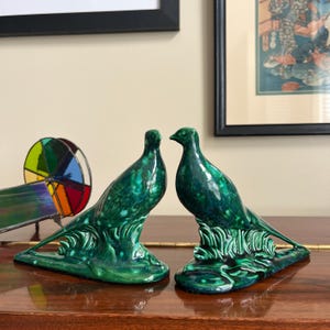 May include: Two green ceramic pheasant figurines with detailed feathers and a leafy base. The figurines are facing each other and are a decorative accent for a home.