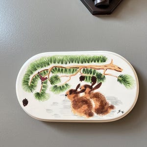 May include: Oval ceramic serving dish with a hand-painted winter scene. The design features two brown rabbits under a pine branch with green needles and a small red bird. The dish has a white background and a cream-colored border.