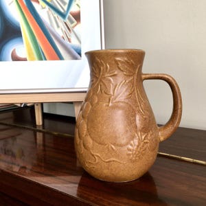 Puede incluir: Una jarra de cerámica marrón con un diseño en relieve de frutas y hojas. La jarra tiene un cuerpo redondeado y un asa.