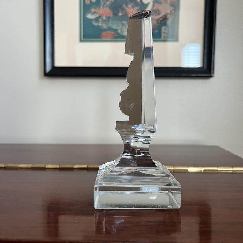 May include: A clear glass decorative object with a faceted design. The object has a square base and a tall, slender top.