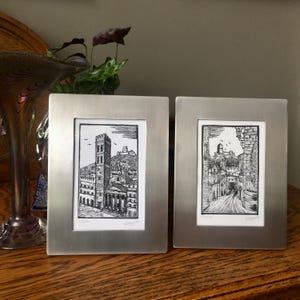 May include: Two black and white framed prints of Italian cityscapes. The prints are in silver frames and are displayed on a wooden surface.